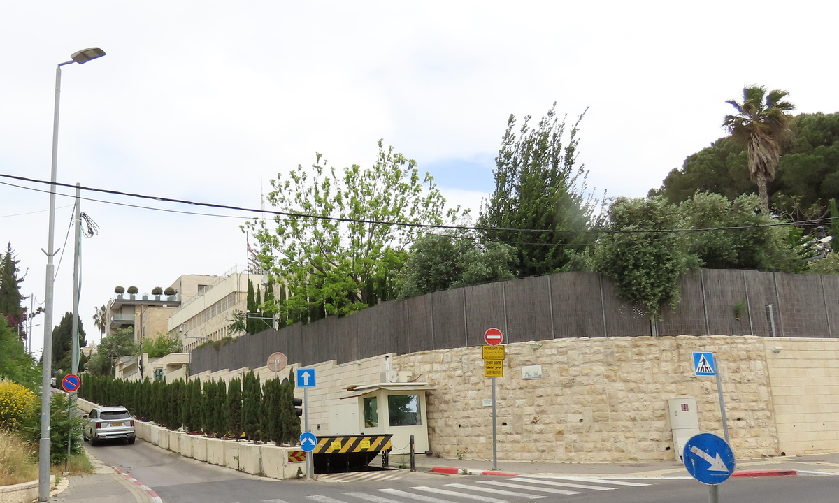 A walled compound on a sloping Jerusalem street: a pale stone wall topped by a dark screen fence, a guard booth and vehicle barrier at the entrance, a multi-storey building behind trees and a small British flag on a pole.