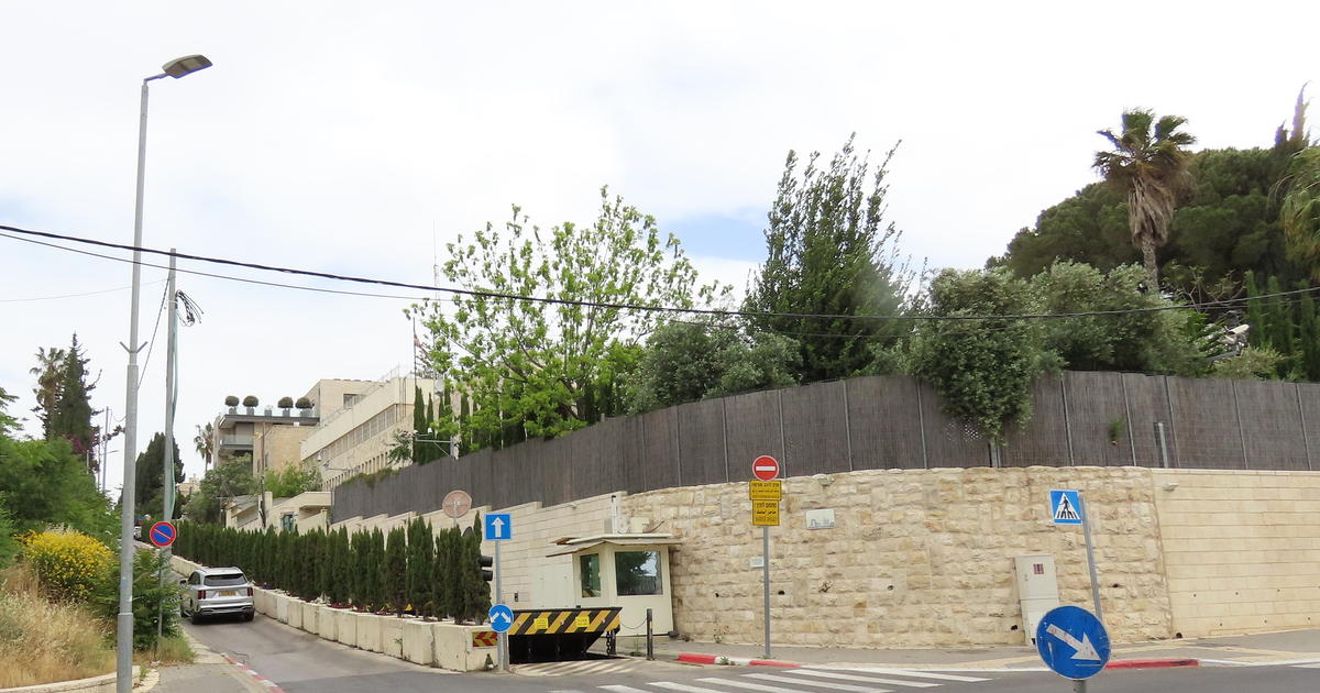 A walled compound on a sloping Jerusalem street: a pale stone wall topped by a dark screen fence, a guard booth and vehicle barrier at the entrance, a multi-storey building behind trees and a small British flag on a pole.