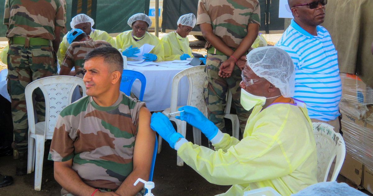A health worker in protective gear and a face shield vaccinates a UN peacekeeper, with other staff in protective suits behind.