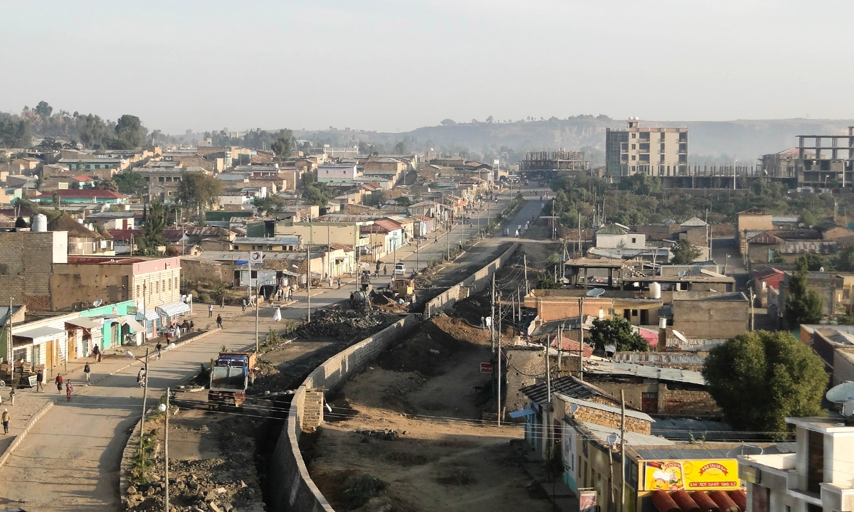 A hazy morning view over Adigrat in November 2012: low buildings line a main road with a truck and a few pedestrians on the left, a walled channel under construction runs through the centre, and taller concrete buildings and hills stand behind.