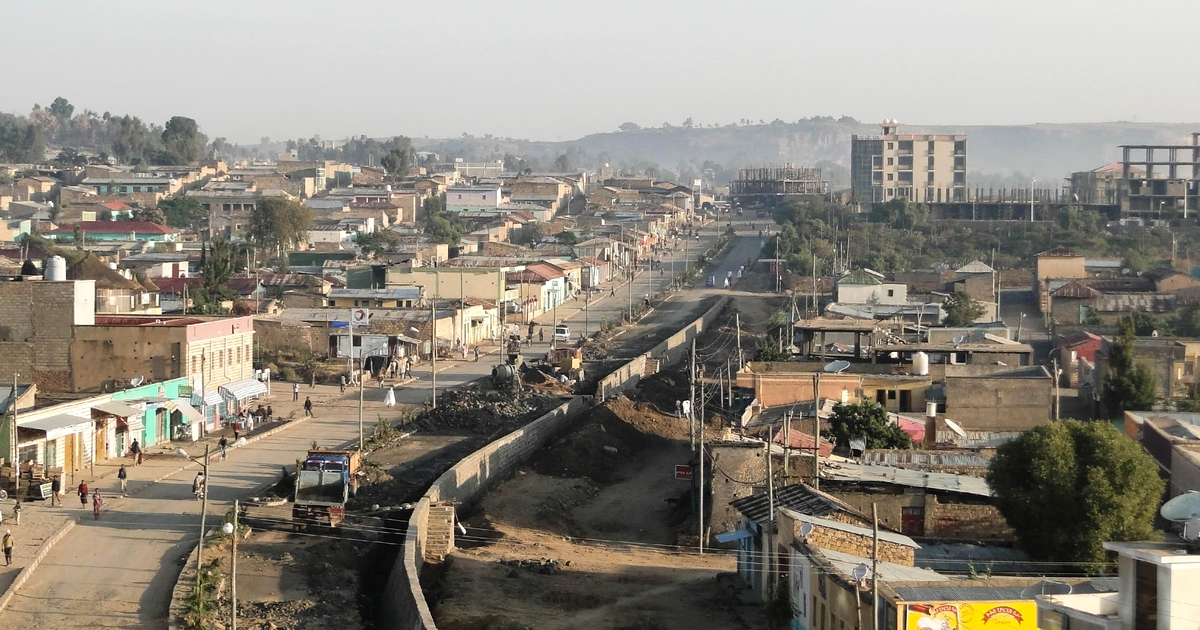 A hazy morning view over Adigrat in November 2012: low buildings line a main road with a truck and a few pedestrians on the left, a walled channel under construction runs through the centre, and taller concrete buildings and hills stand behind.