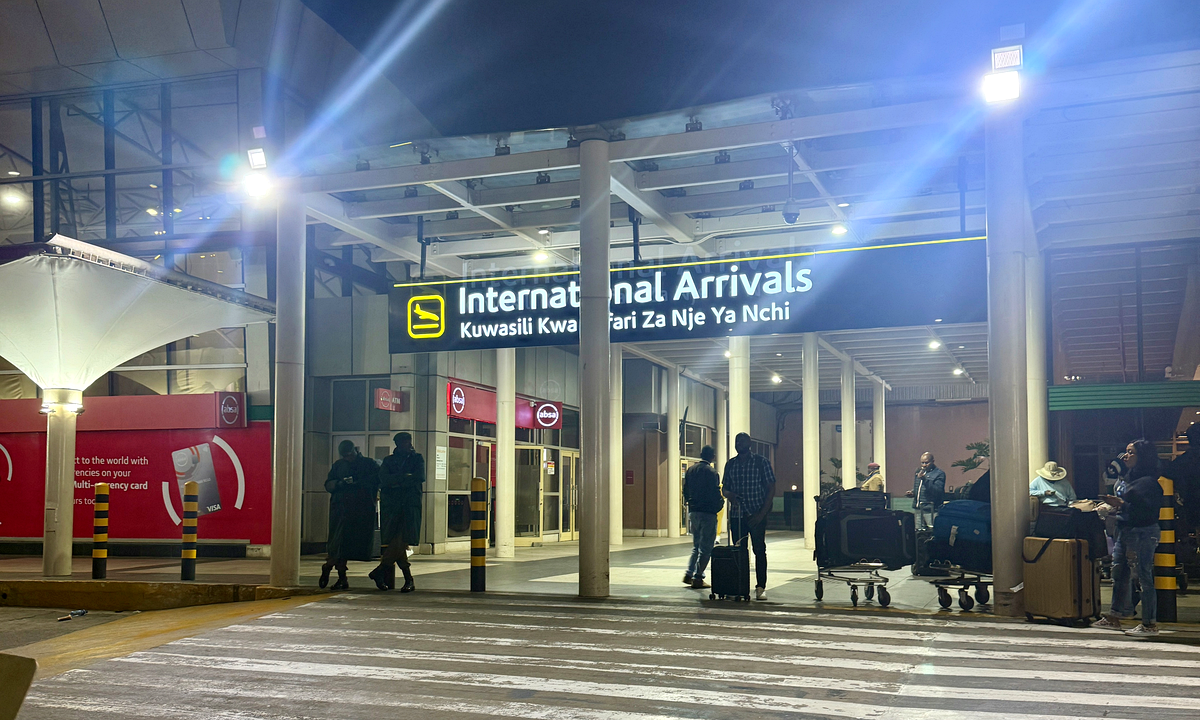 The international arrivals entrance at Jomo Kenyatta International Airport in Nairobi at night, under a lit sign reading International Arrivals in English and Swahili, with travellers and luggage trolleys. Photograph: Fawaz.tairou / Wikimedia Commons, CC BY 4.0.