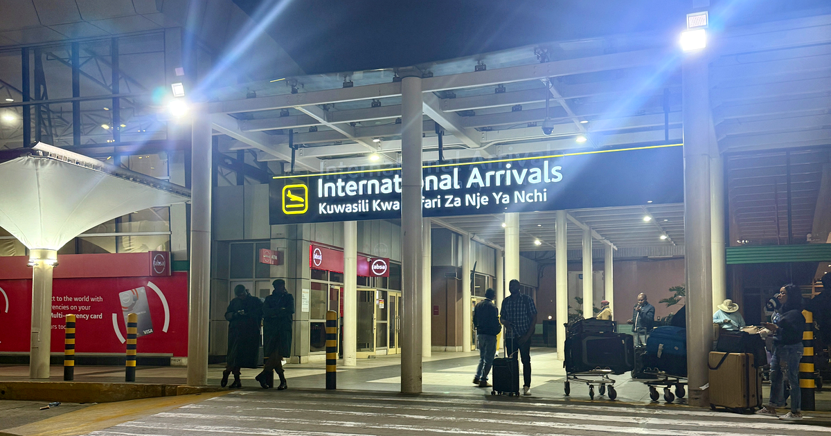 The international arrivals entrance at Jomo Kenyatta International Airport in Nairobi at night, under a lit sign reading International Arrivals in English and Swahili, with travellers and luggage trolleys. Photograph: Fawaz.tairou / Wikimedia Commons, CC BY 4.0.