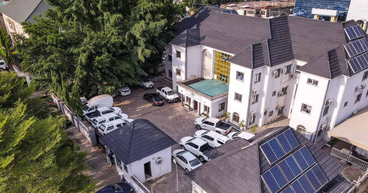 Aerial view of the Nigeria Centre for Disease Control and Prevention headquarters in Abuja: white three-storey buildings with grey roofs and solar panels, a car park of white vehicles and trees beside the entrance. Photo from 2021.