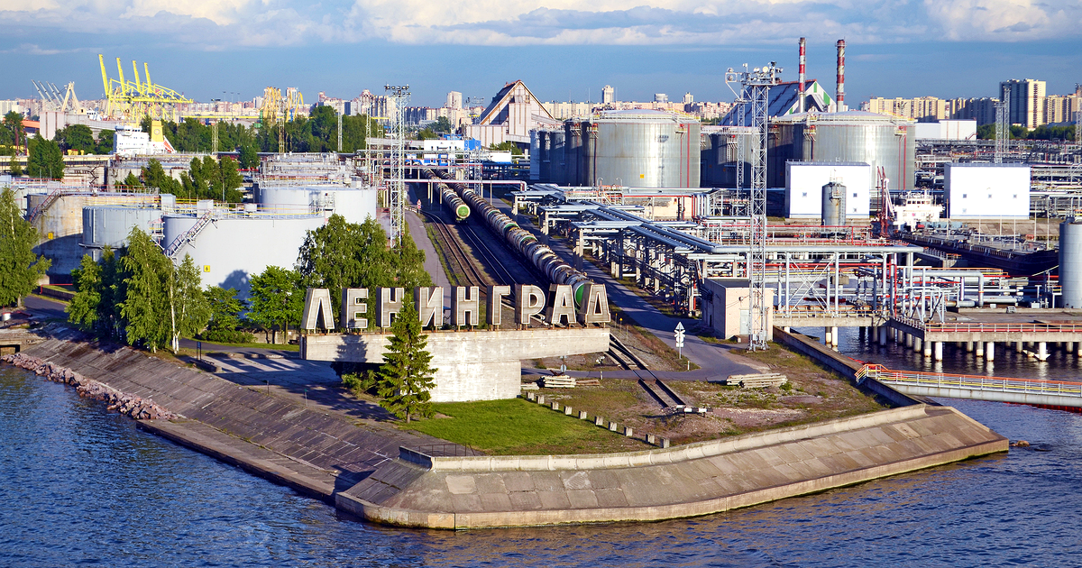 Archive view from the water of oil storage tanks, rail tank cars and loading gantries behind a large concrete sign reading ЛЕНИНГРАД on a stone pier.