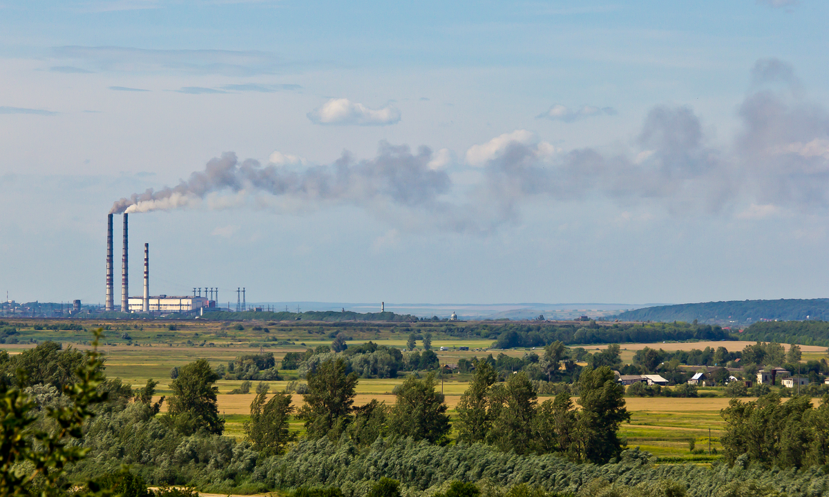 The Burshtyn coal-fired power station in western Ukraine: three tall chimneys on a flat horizon venting a long grey plume across a pale summer sky, with green fields, trees and village houses in the foreground.