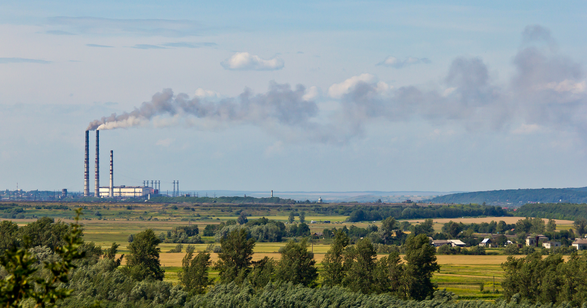 The Burshtyn coal-fired power station in western Ukraine: three tall chimneys on a flat horizon venting a long grey plume across a pale summer sky, with green fields, trees and village houses in the foreground.