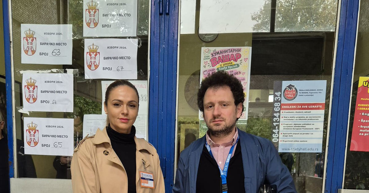 Two international election observers wearing accreditation badges stand at the glass doors of a Belgrade school, where official signs bearing Serbia's coat of arms read "Elections 2026, polling station" with station numbers including 63, 64, 65 and 67.