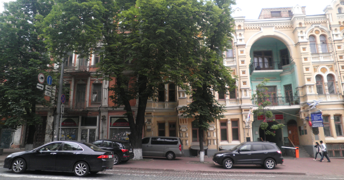 A row of ornate older buildings on a Kyiv street in May 2012, with a cream facade and arched balcony on the right, flags at the entrance, trees and parked cars in front.