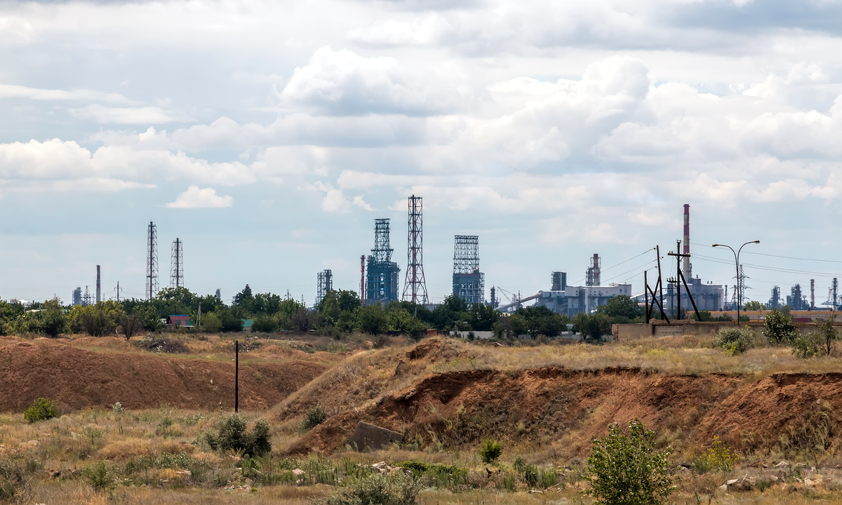 Distillation towers, steel lattice towers and a tall red-and-white chimney of an oil refinery on the horizon under a cloudy sky, beyond dry grassland and eroded orange earth banks in the foreground.