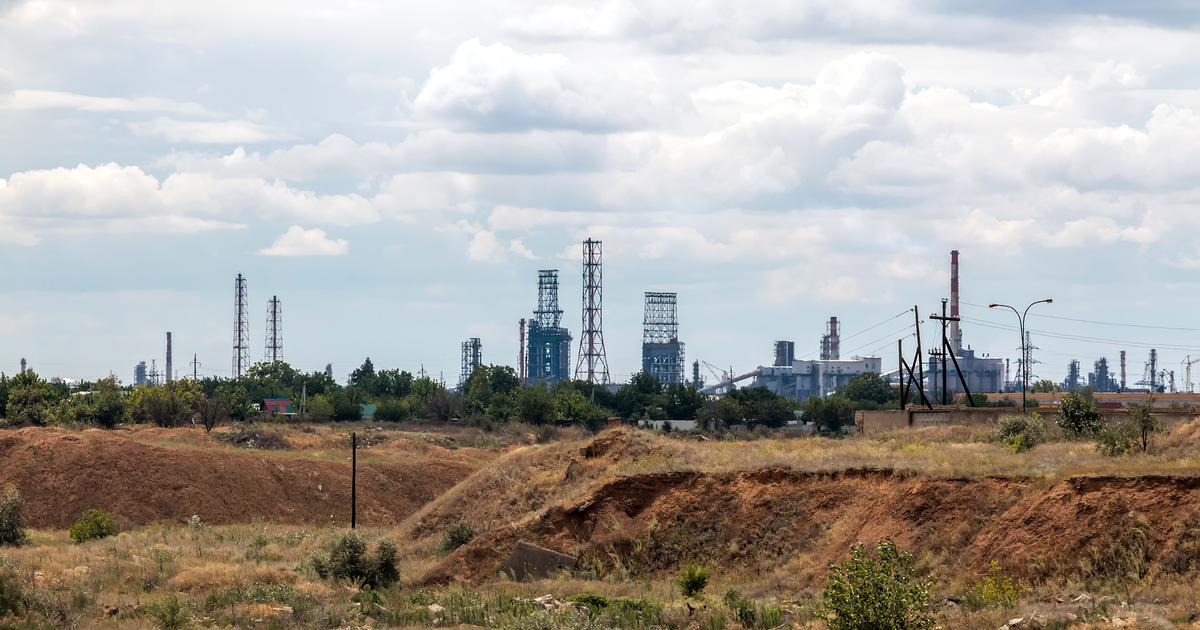 Distillation towers, steel lattice towers and a tall red-and-white chimney of an oil refinery on the horizon under a cloudy sky, beyond dry grassland and eroded orange earth banks in the foreground.